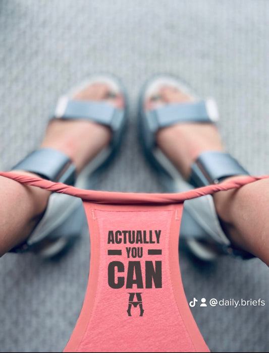 Pink seamless comfy underwear with 'Actually You Can' text worn by a person in sandals on a gray floor.