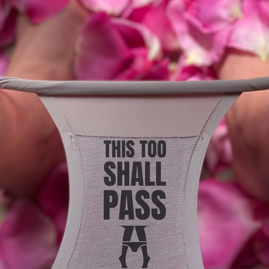  'THIS TOO SHALL PASS' text and logo, held against a pink floral background.